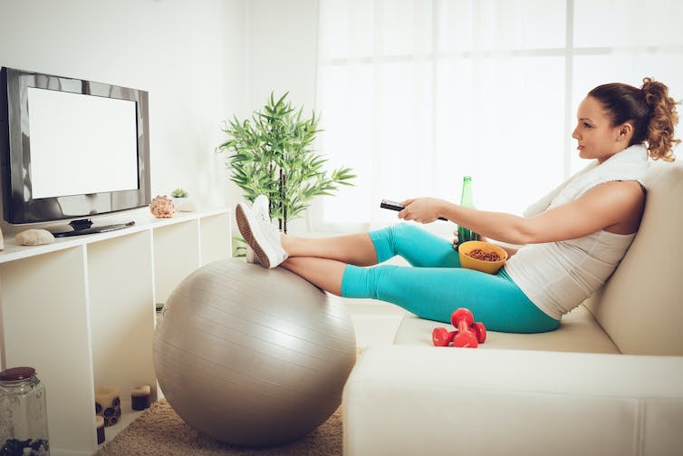 A woman in fitness clothes sitting on a sofa snacking and ignoring her fitness equipment while turning on the tv